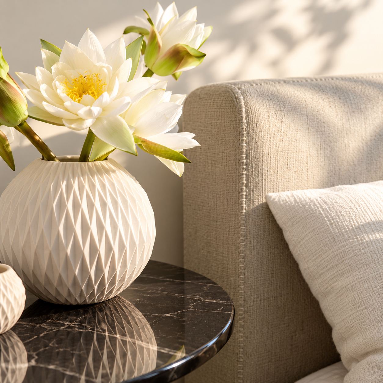 White water lilies in a faceted ceramic vase on a dark marble side table beside an upholstered chair